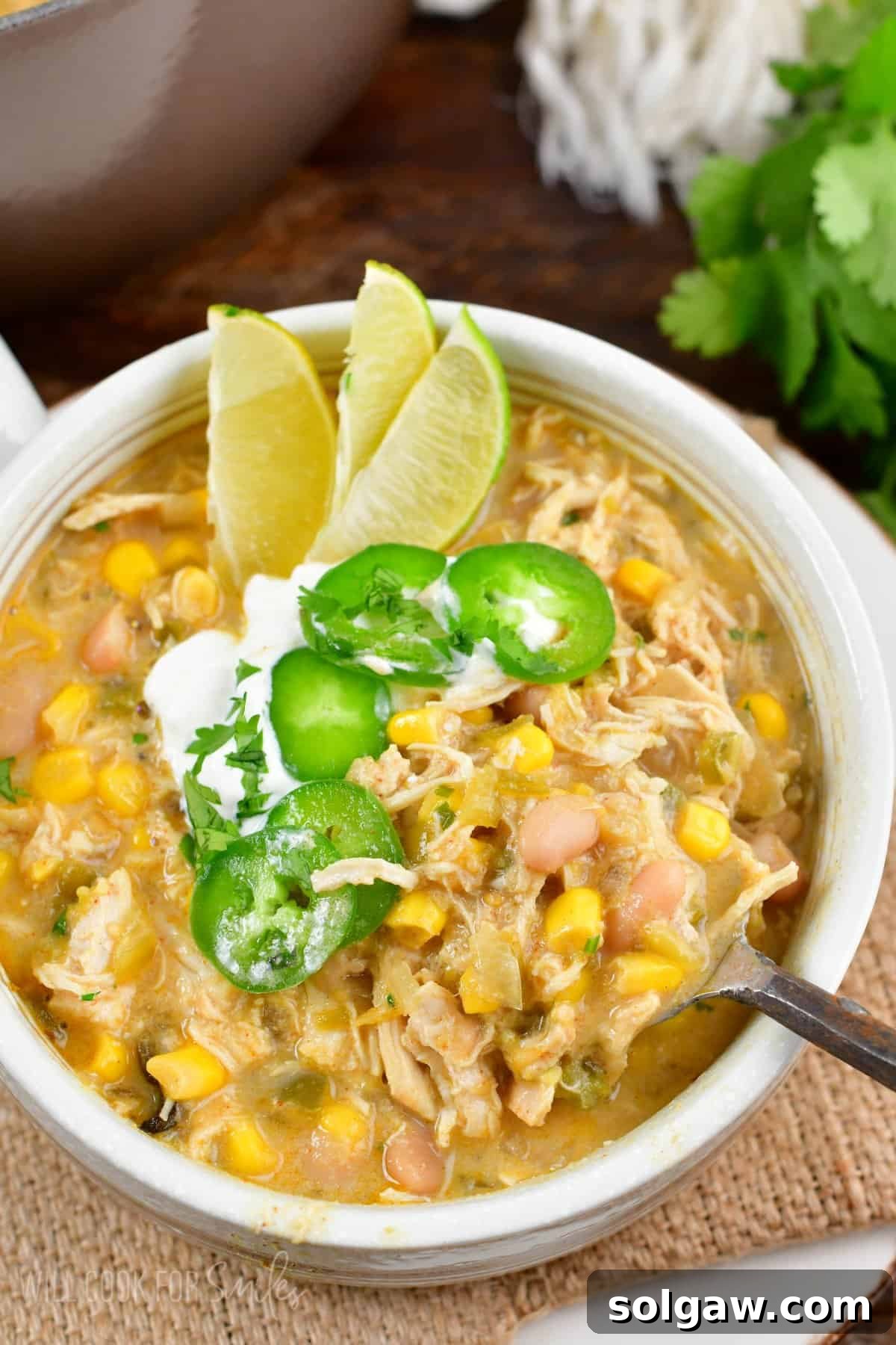 Creamy White Chicken Chili 6 white chicken chili in a bowl with three sliced limes, jalapeno, and sour cream on top with a spoon scooping some out,