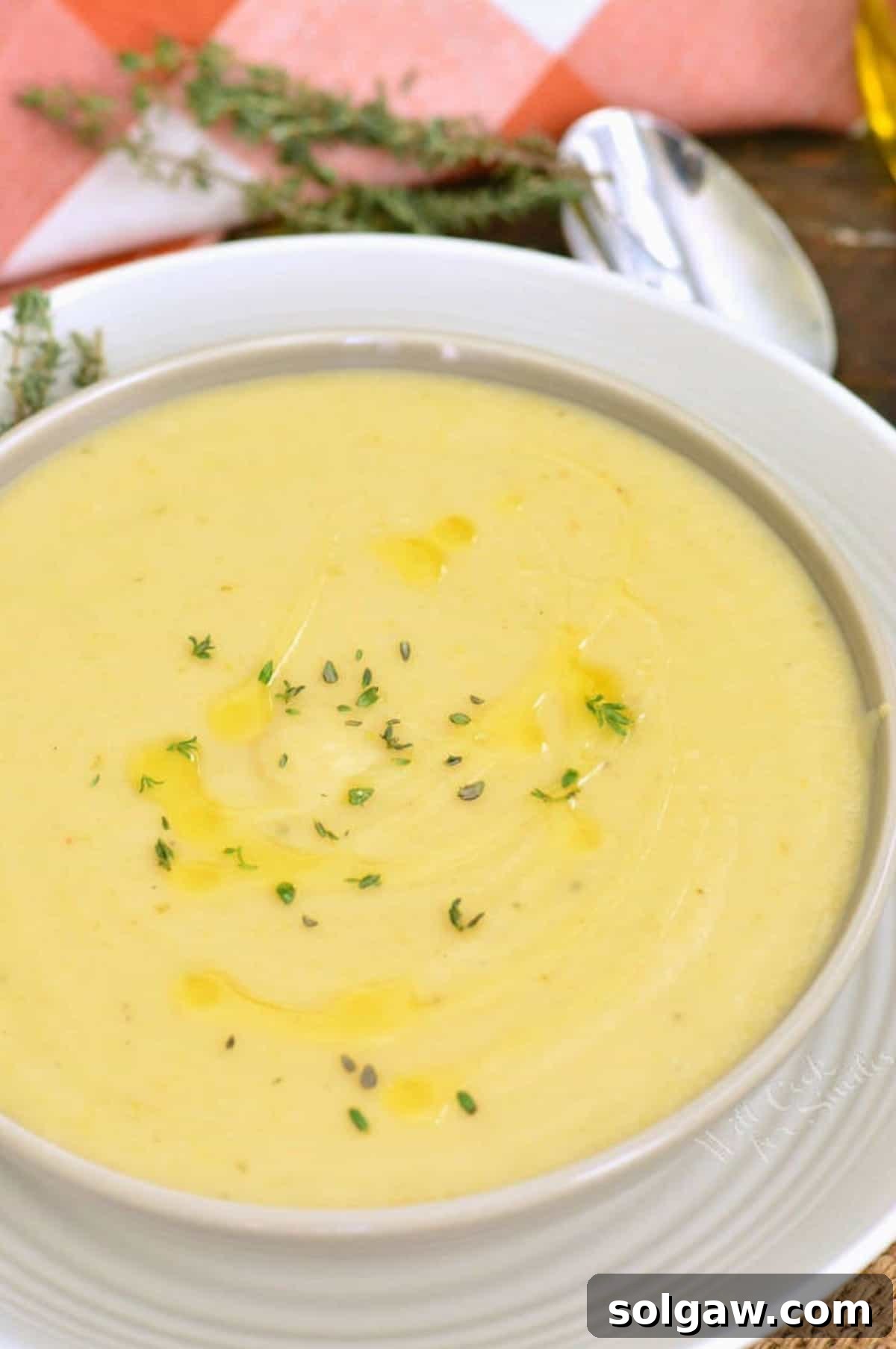 A deep bowl of potato leek soup placed on a white plate, garnished with fresh rosemary sprigs.