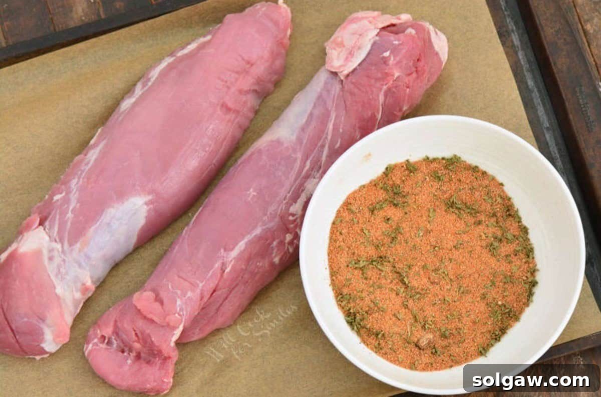 two pork tenderloins uncooked on a parchment lined baking sheet with a bowl of seasoning next to it.