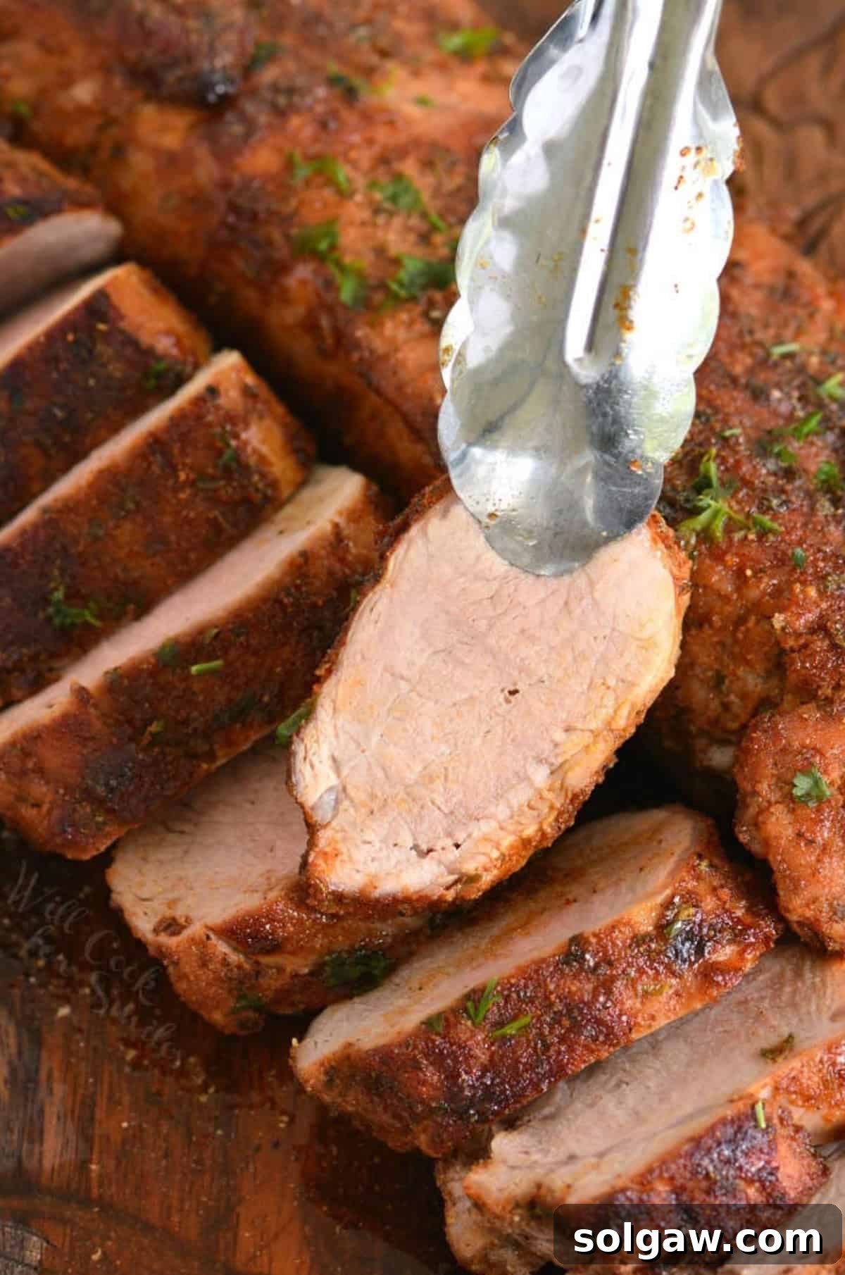 Two roasted pork tenderloins one sliced and one unsliced holding a slice with a pair of tongs.