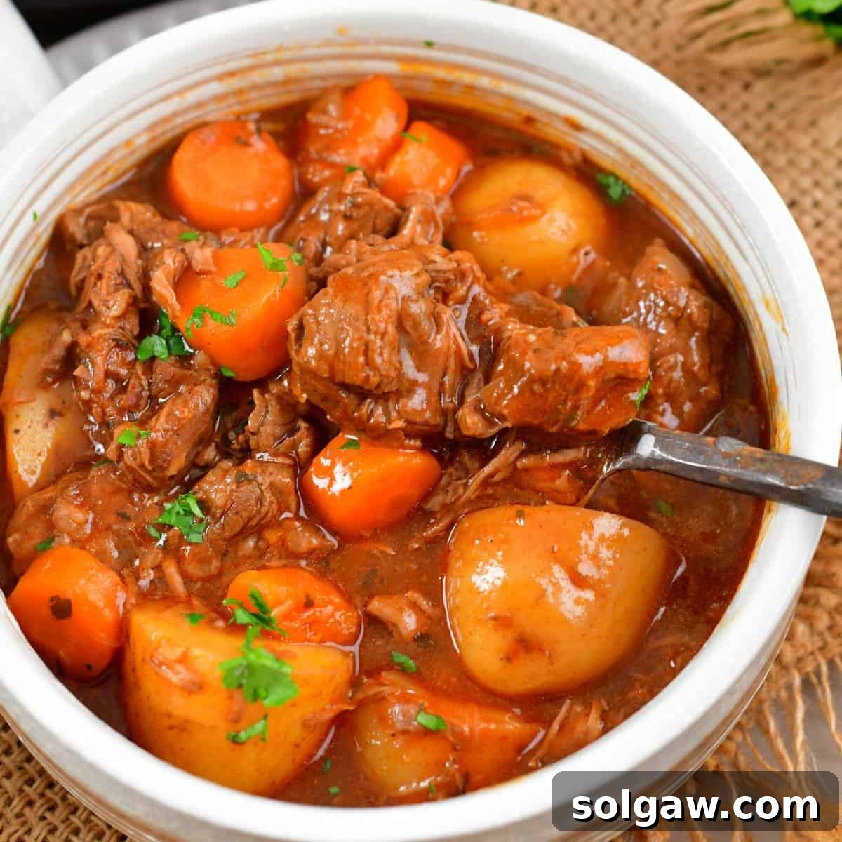 A light bowl filled with beef stew and vegetables and a silver spoon.
