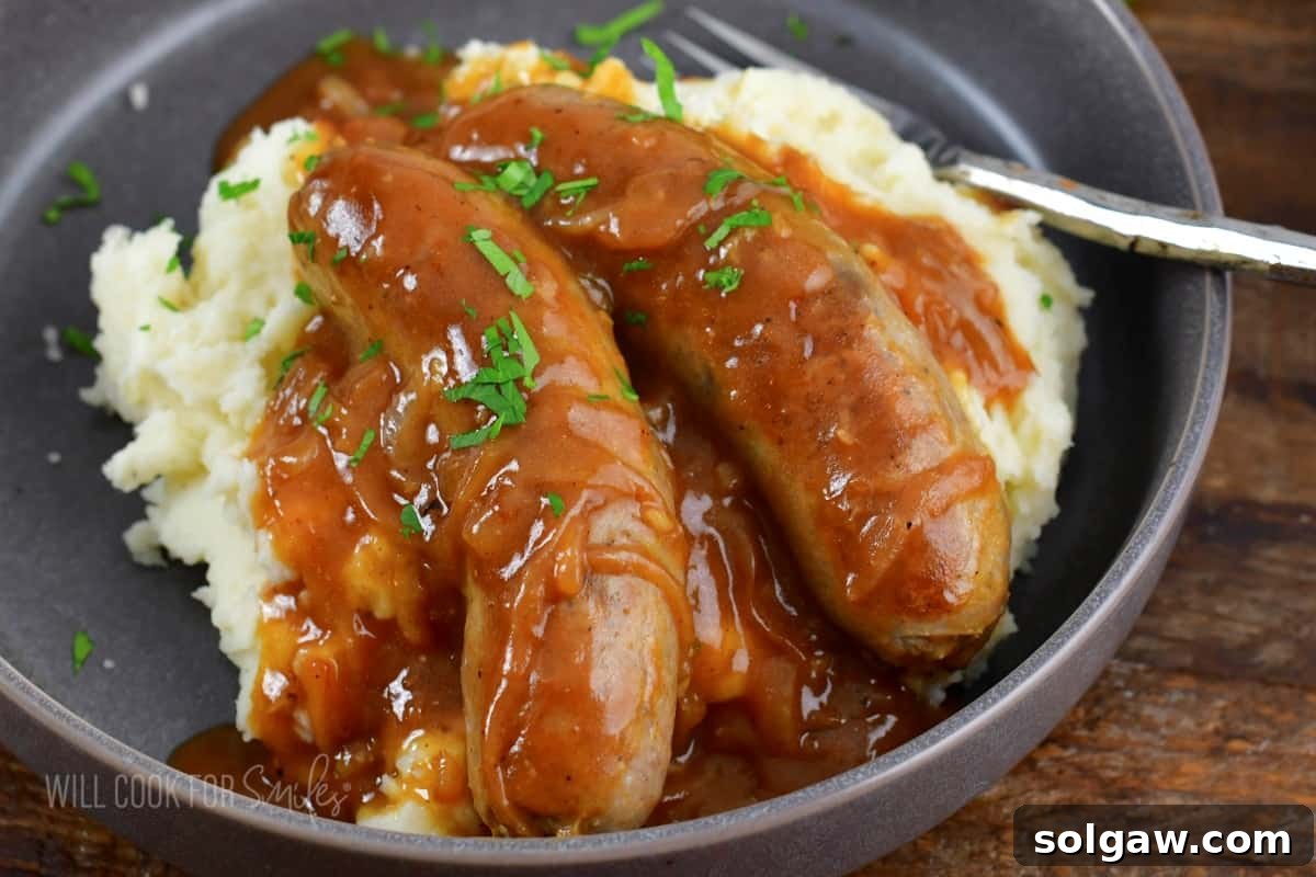 Onion Gravy Smothered Sausages and Mash 6 sausages in onion gravy over mashed potatoes in a plate.