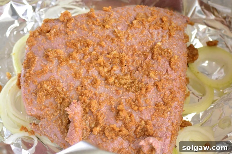 One-Pan Corned Beef with Crispy Potatoes 5 Oven-baked corned beef brisket generously coated with mustard rub, placed on a bed of sliced onions in a roasting pan, ready for the oven.