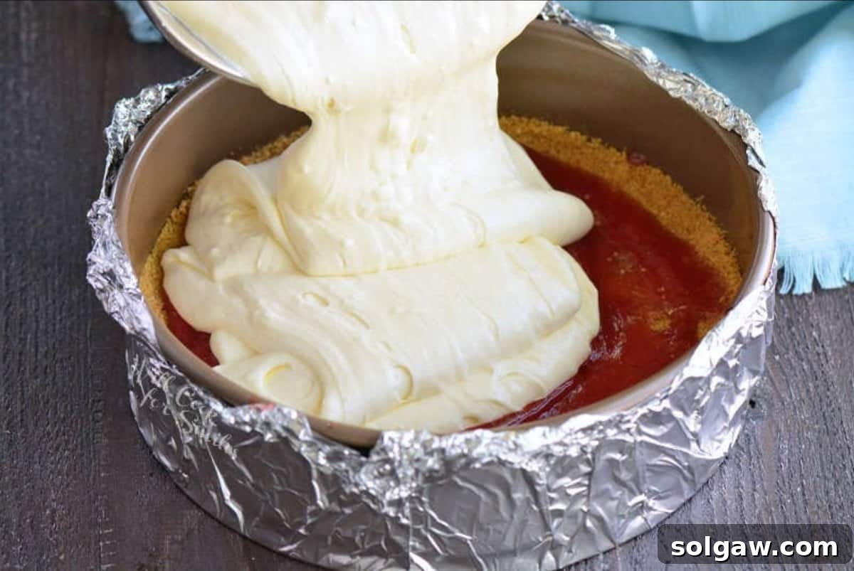 Cheesecake batter being poured over a prepared crust with a layer of strawberry sauce in a springform pan.