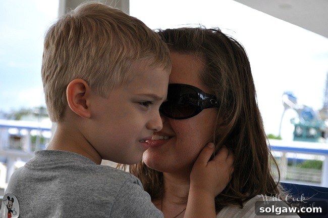 A heartwarming image of a mother and her young son smiling happily together, enjoying a ride on the PeopleMover at Disney World, with the vibrant park scenery blurred in the background, capturing a moment of pure family joy.