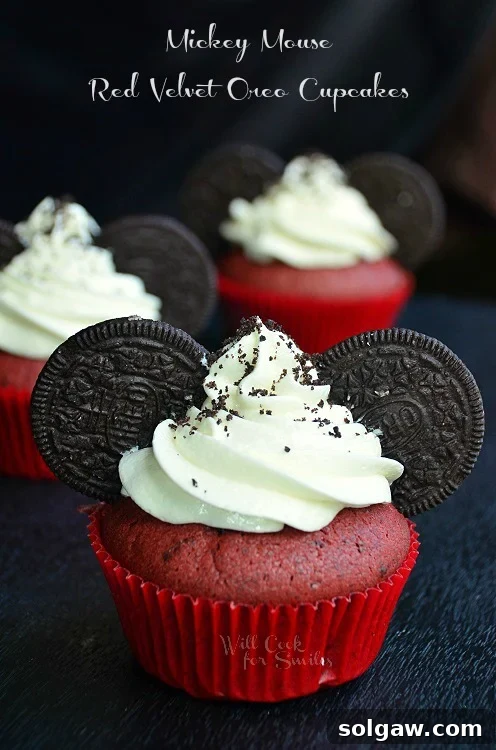 A stunning close-up, vertical shot of a single Mickey Mouse Red Velvet Cupcake, showcasing its rich, vibrant red cake beneath a generous swirl of pristine white cream cheese frosting and charming Oreo 'ears'. The image emphasizes the beautiful texture and festive appeal of the cupcake.