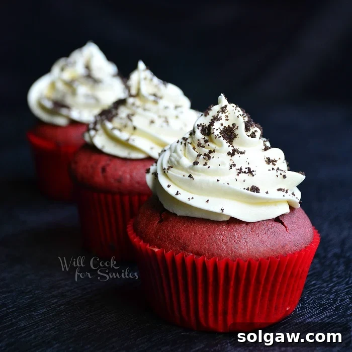Three perfectly crafted Red Velvet Oreo Cupcakes, each topped with a generous swirl of white cream cheese frosting and a scattering of crushed Oreos, artfully arranged on a sleek black surface, highlighting their rich colors and inviting textures.
