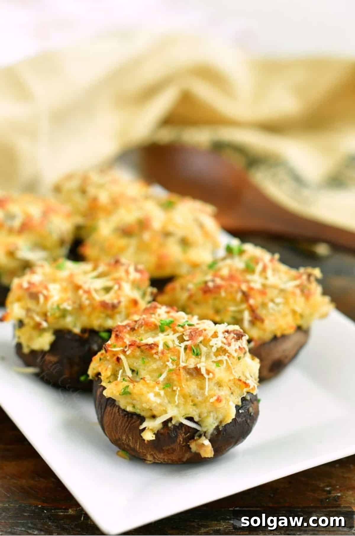 Several golden-brown crab stuffed mushrooms arranged on a white plate, with a wooden spoon and burlap towel in the soft-focus background, highlighting their appetizer appeal.