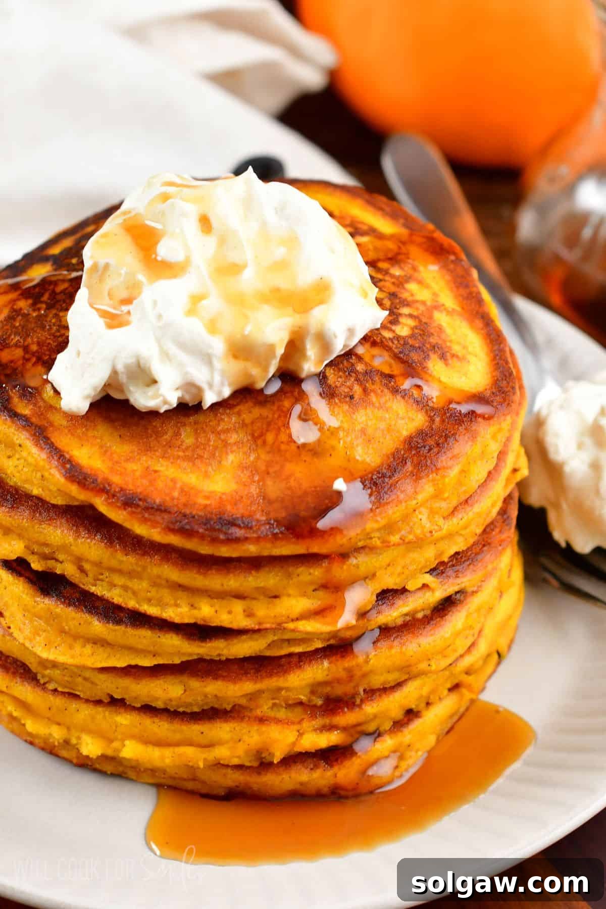 A stack of pumpkin pancakes with whipped cream and syrup on top on a plate, ready for a delicious fall breakfast.