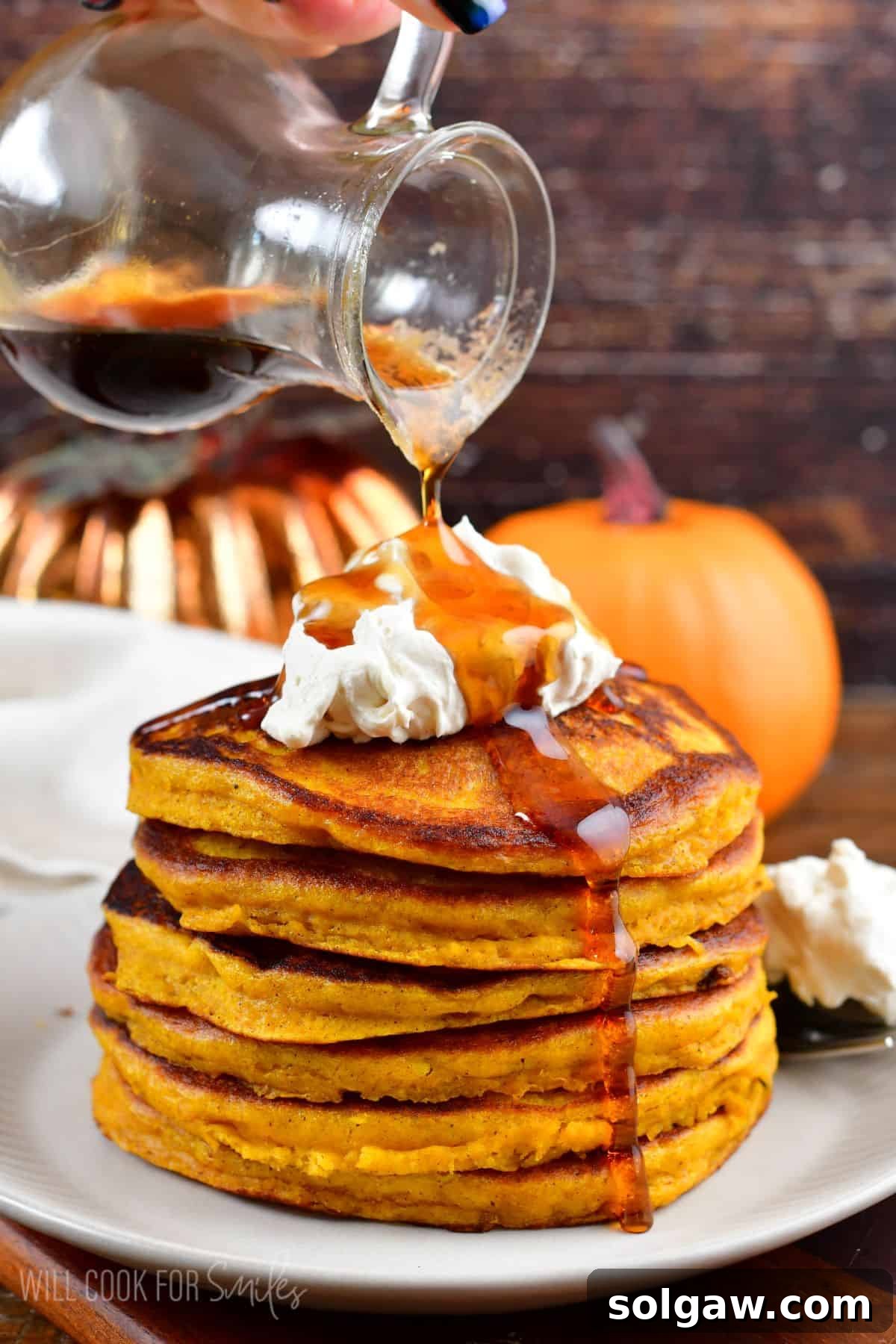 A stack of golden pumpkin pancakes topped with whipped cream, with maple syrup being poured over them, creating a mouth-watering visual.