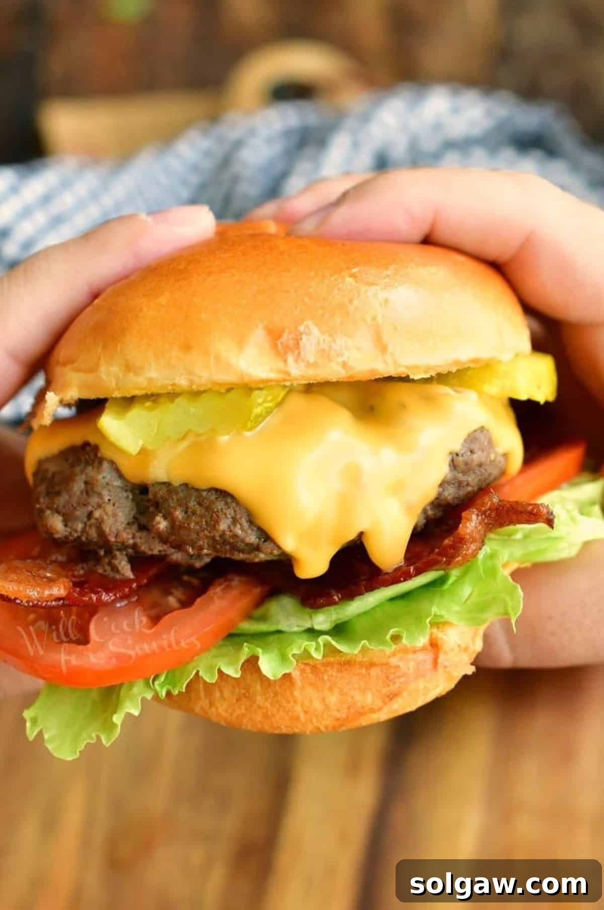 holding a cheeseburger with tomatoes lettuce and pickles with two hands.