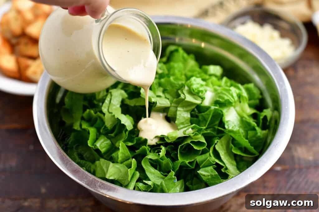 Pouring creamy white Caesar salad dressing over crisp Romaine lettuce leaves in a clear salad bowl