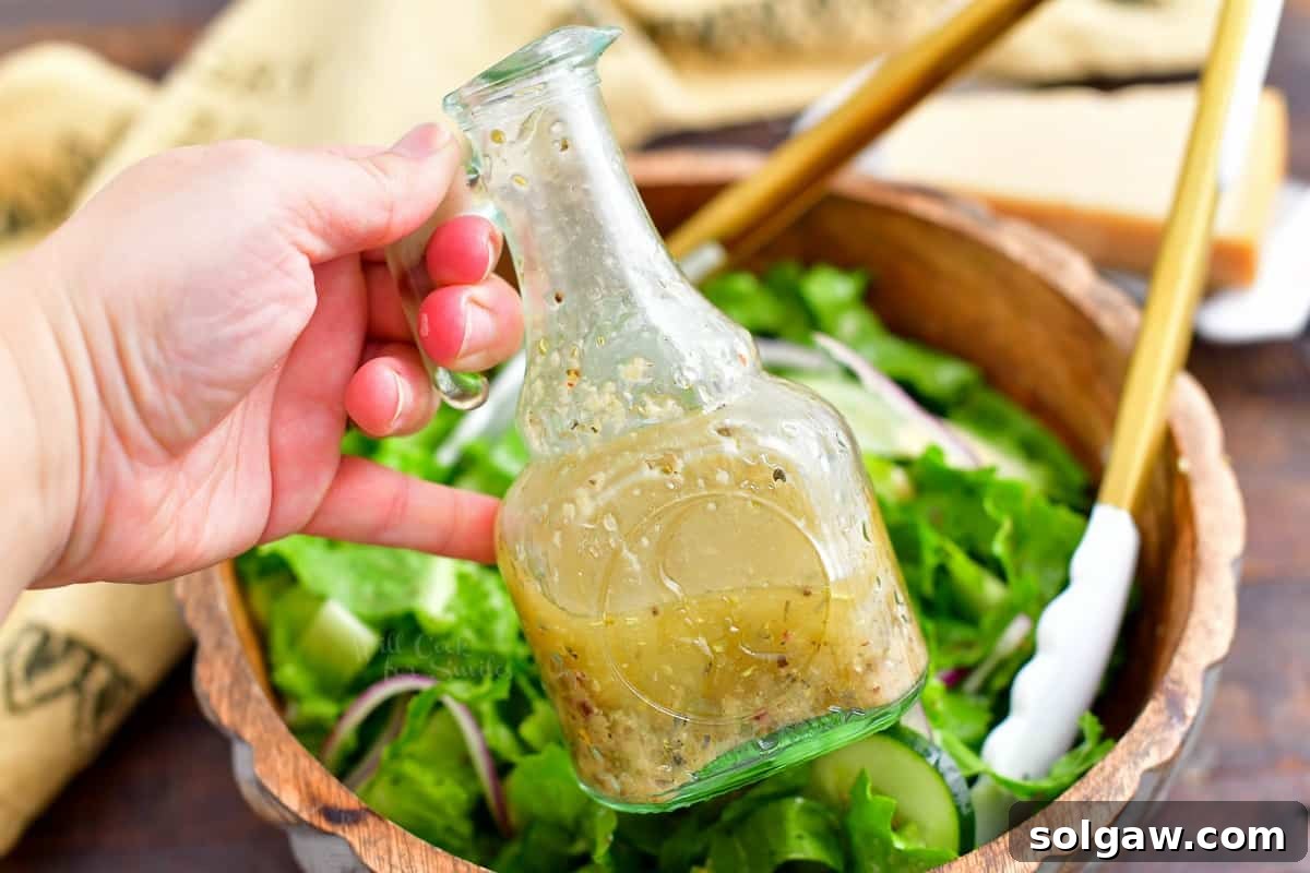 Zesty Italian Vinaigrette 3 closeup: woman's hand holding bottle of homemade Italian dressing; bowl of salad in background, emphasizing fresh ingredients.