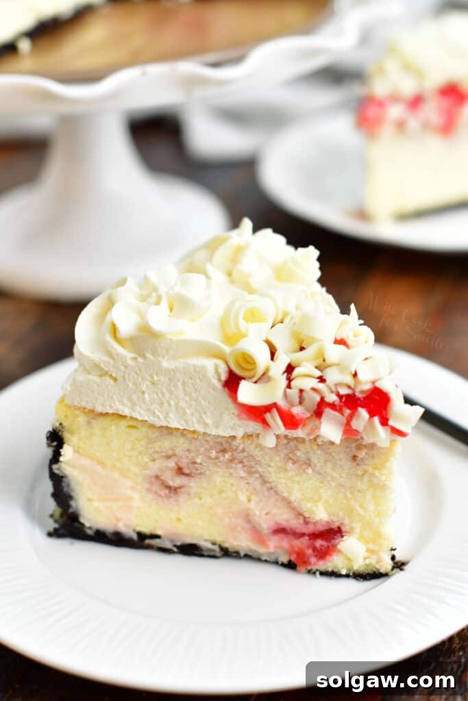 A perfectly sliced piece of white chocolate raspberry cheesecake, topped with fluffy cream cheese frosting, a drizzle of crimson raspberry sauce, and delicate white chocolate curls, elegantly presented on a plate with the full cheesecake on a stand in the background.
