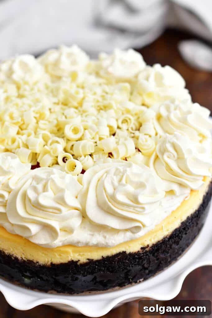 A whole, beautifully decorated white chocolate raspberry cheesecake, showcasing its dark Oreo cookie crust, smooth white frosting, and elegant white chocolate shavings on top, resting on a rustic wooden surface.