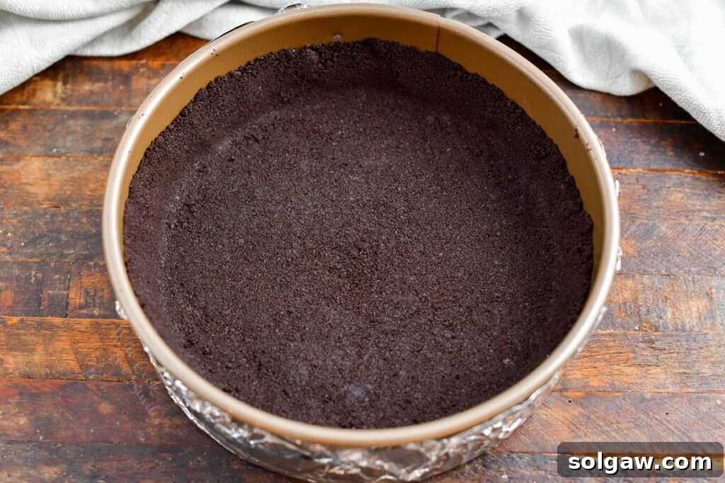 Close-up of a chocolate cookie crust being pressed into a springform pan, ready for the cheesecake filling, illustrating the first step in the recipe.
