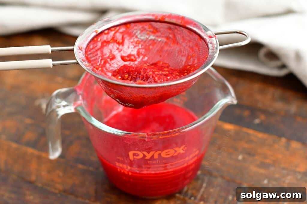 Hands pressing raspberries through a fine-mesh strainer to create a smooth raspberry puree, a crucial step for the vibrant raspberry swirl in the cheesecake.