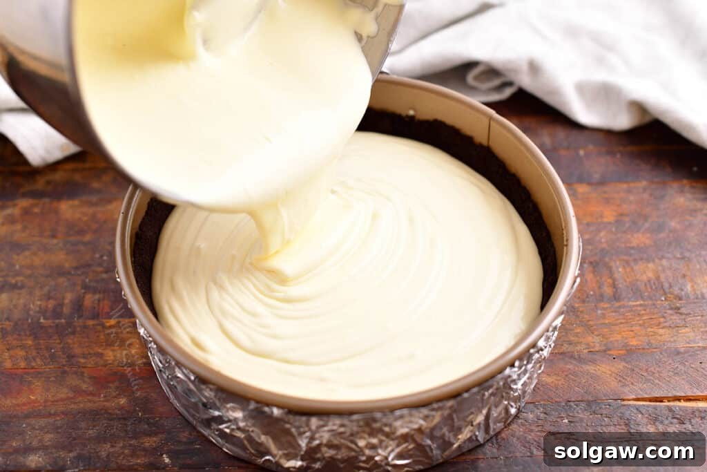 Creamy white chocolate cheesecake filling being poured over a dark chocolate cookie crust in a springform pan, surrounded by foil for a water bath.