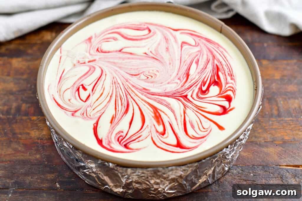 A mesmerizing swirl of white chocolate cheesecake filling and vibrant raspberry sauce within a foil-wrapped springform pan, poised for baking in a water bath.