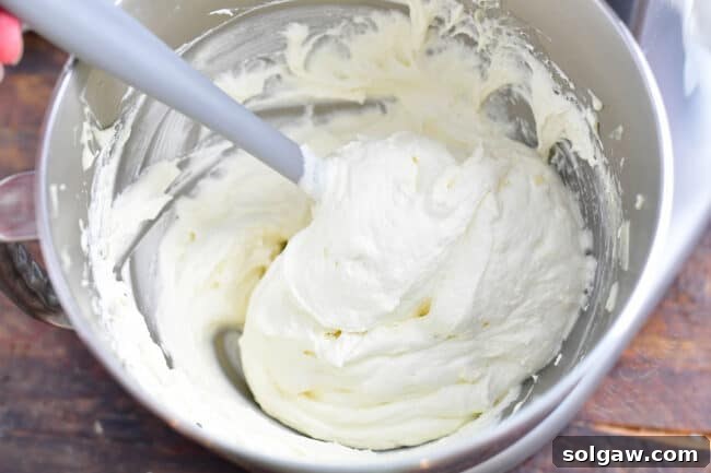 Close-up view of fluffy cream cheese frosting being gently mixed in a large white bowl with a spatula, illustrating the preparation of the topping for the cheesecake.