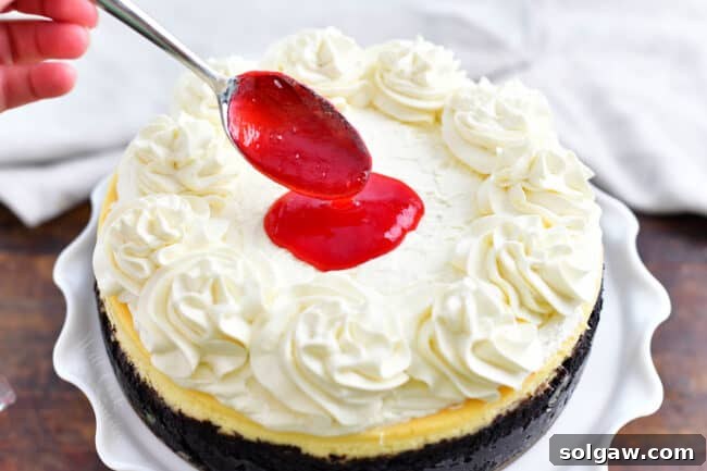 A spoonful of vibrant raspberry sauce being drizzled over the smooth white top of a white chocolate cheesecake with an Oreo crust, ready for further decoration.