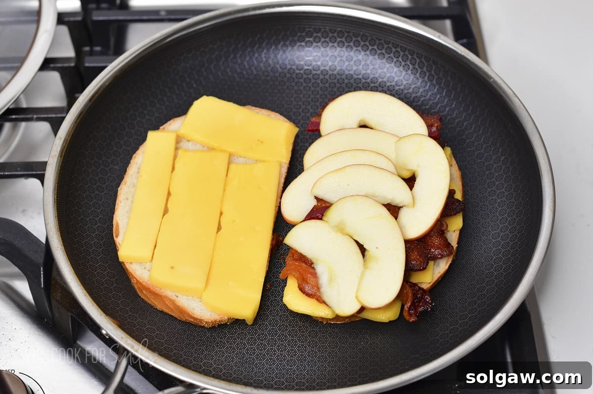 Two slices of bread in a pan with cheese, one of which is topped with bacon and thinly sliced apple, ready to be combined.