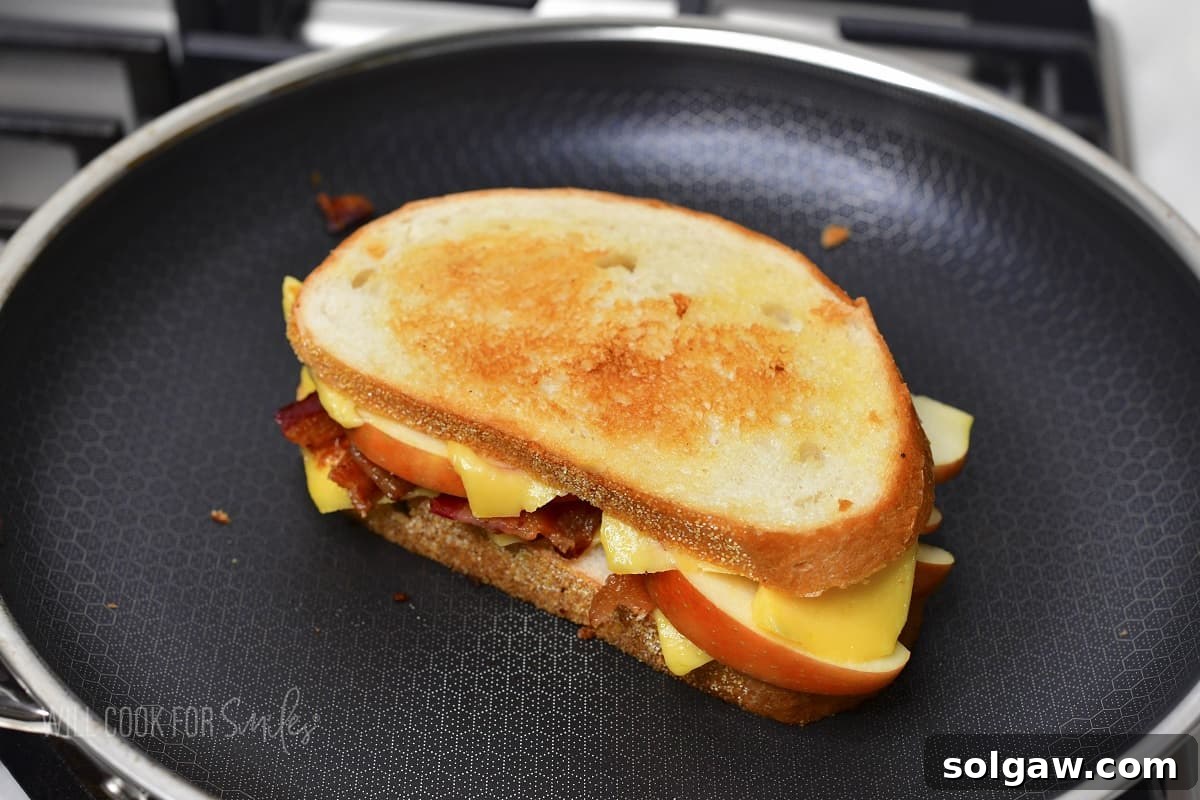 A grilled cheese sandwich with bacon and apple cooking in a hot pan, with melted cheese oozing slightly.