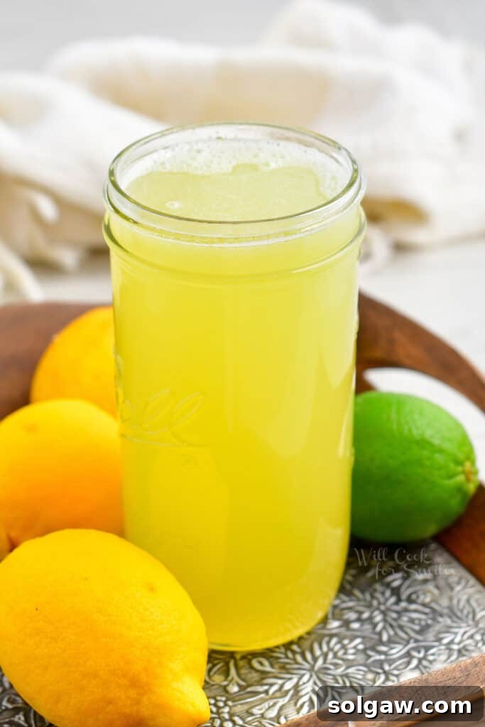 Freshly made sweet and sour mix in a clear glass jar, surrounded by whole lemons and limes on a rustic wooden surface. The liquid is a bright, clear yellow, inviting and refreshing.