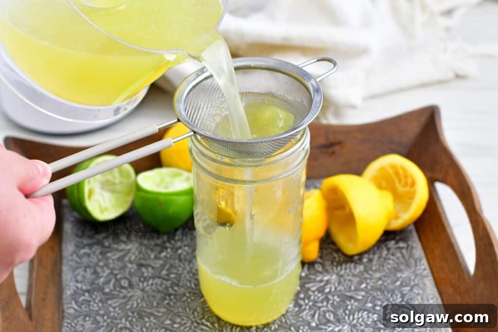 A fine mesh sieve held over a tall glass jar, with homemade cocktail mixer being poured through it, demonstrating the straining process to remove pulp and ensure a smooth texture.
