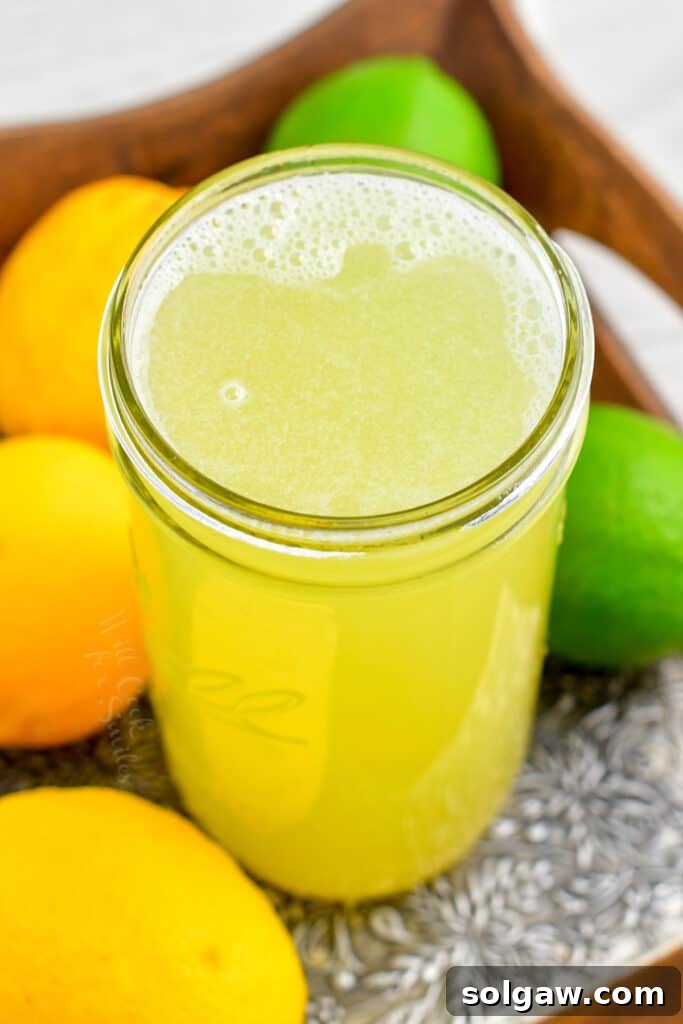 An overhead shot of a large glass jar filled with homemade sweet and sour mix, surrounded by fresh lemons and limes, indicating readiness for use in cocktails. The setup is clean and inviting.