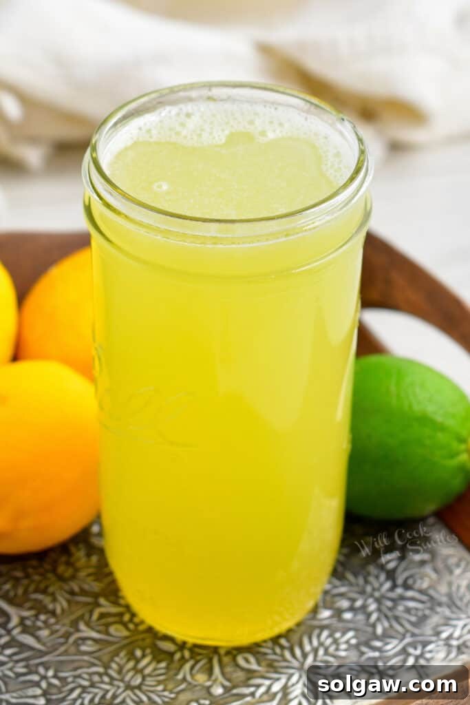 A tall, elegant glass jar filled with golden homemade sweet and sour mix, positioned centrally on a wooden board. It is surrounded by an abundance of fresh, ripe lemons and limes, highlighting the natural ingredients.