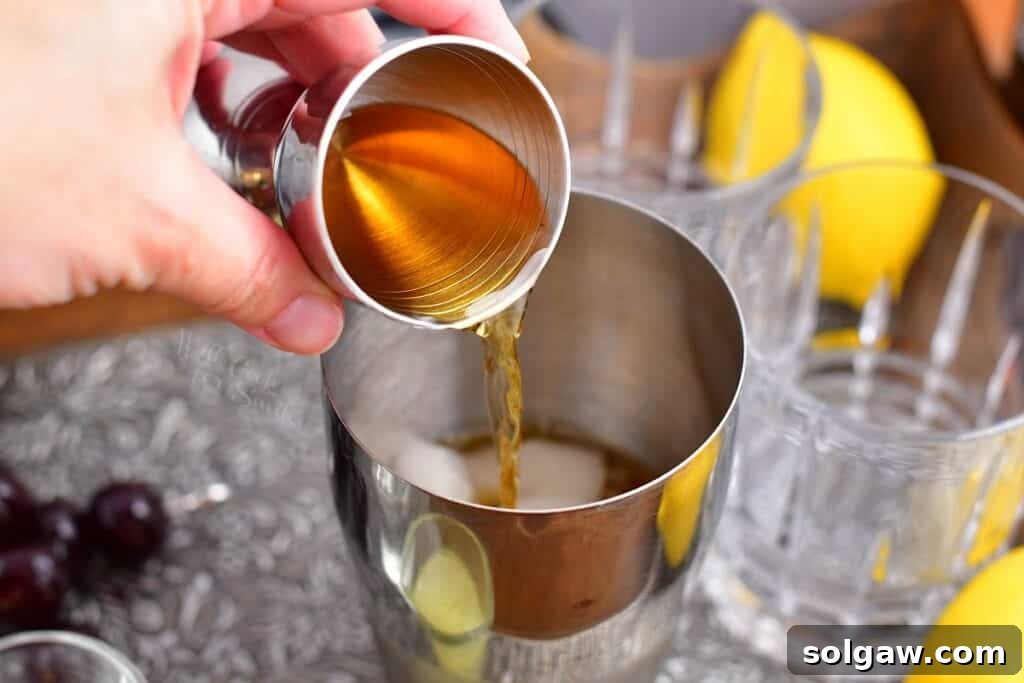 Velvet Almond Sour 5 A lighter liquid, possibly bourbon, is being poured into the cocktail shaker containing amaretto and ice.