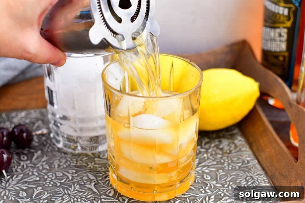 Velvet Almond Sour 7 An Amaretto Sour, freshly shaken, is being poured through a cocktail strainer into a glass half filled with ice.