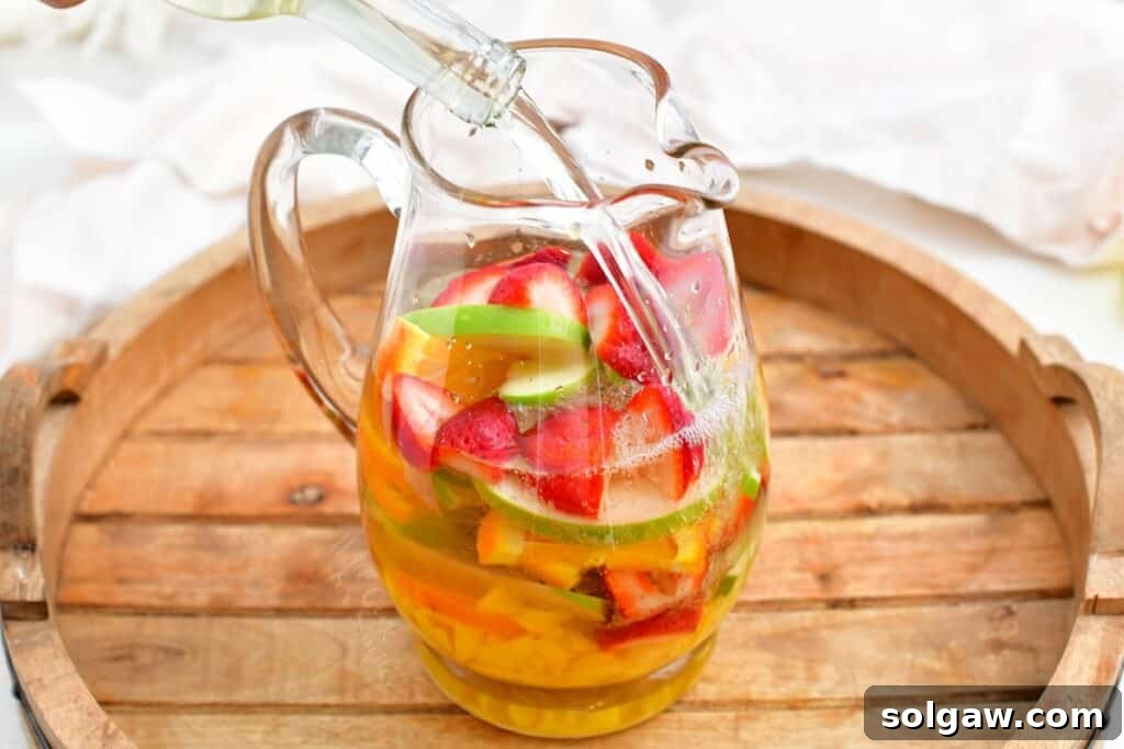 Summer's Perfect White Sangria 4 Clear white wine and orange liqueur are being poured into a large glass pitcher filled with sliced fresh fruit, ready to infuse.