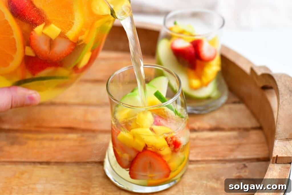 Summer's Perfect White Sangria 6 A pitcher of white sangria is pouring its contents, including sliced fruits, into a small, elegant round glass, ready for enjoyment.