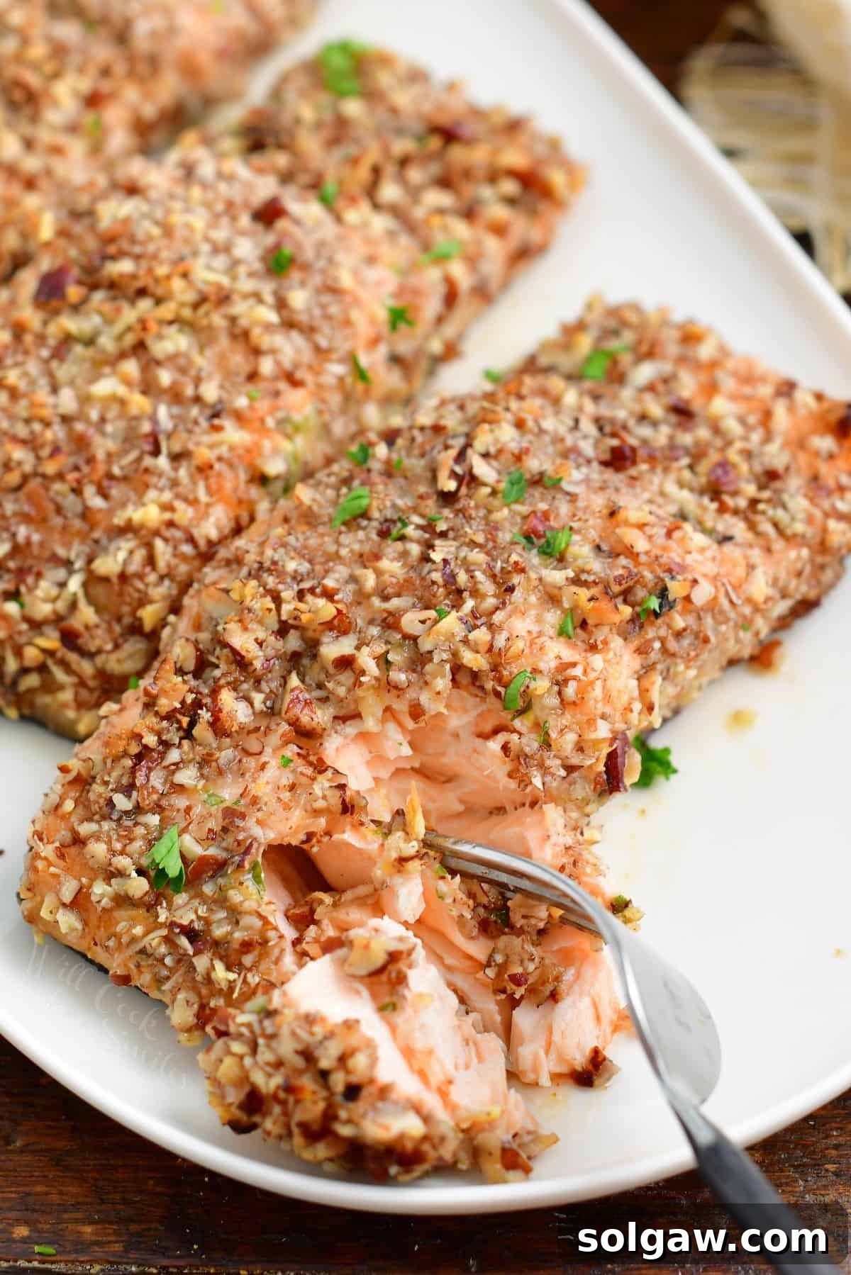 A fork is removing a bite-sized piece of pecan crusted salmon from a white plate, showcasing its golden crust and flaky interior.