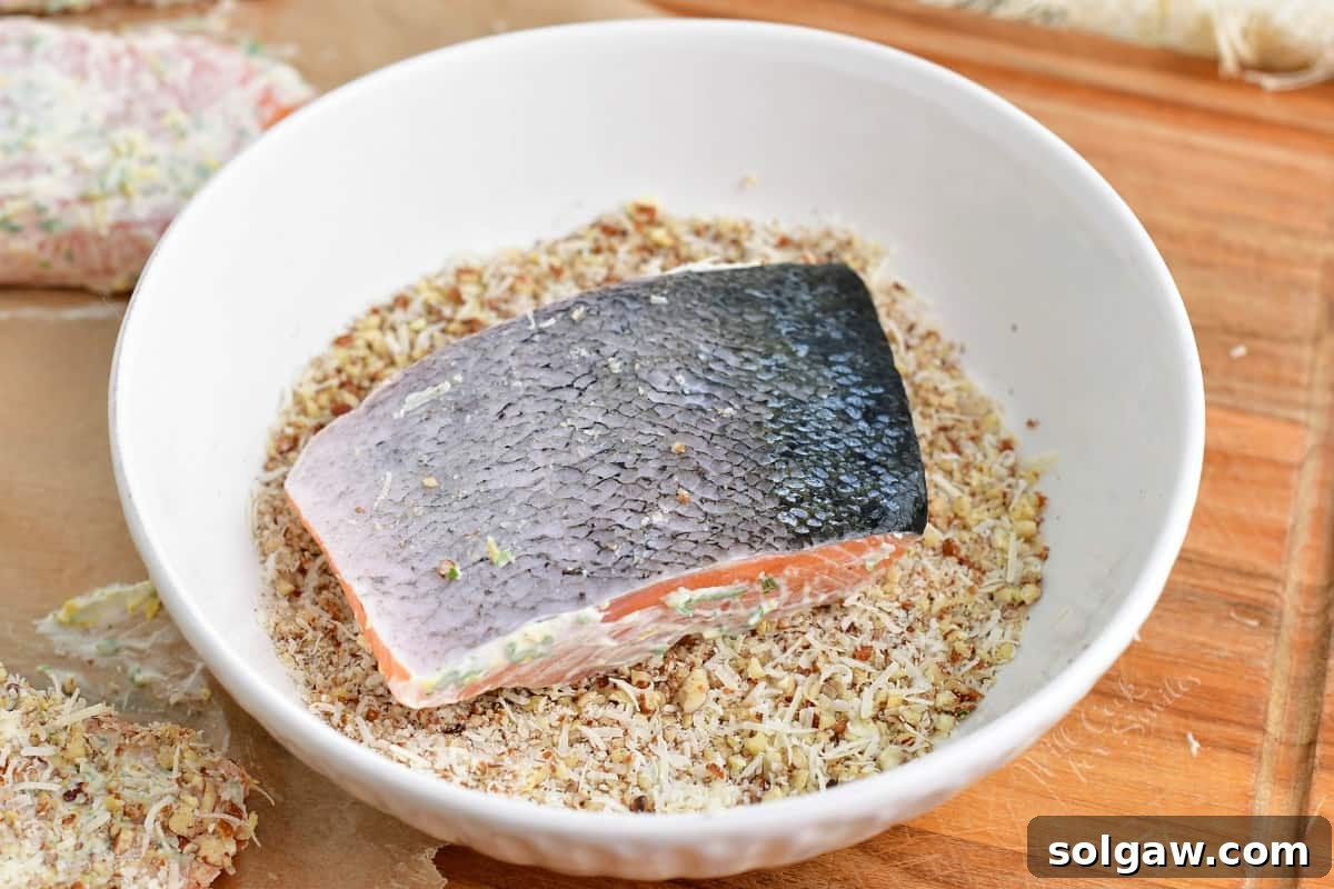 A salmon fillet is carefully dipped into a bowl of crushed pecans and Parmesan, ensuring an even coating for the crust.