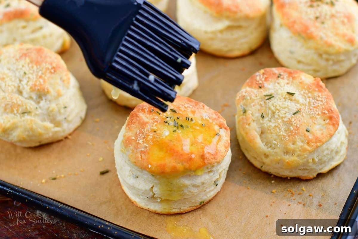 Parmesan Herb Garlic Butter Biscuits 8 A hand brushing melted garlic butter onto a warm, baked biscuit on a baking sheet.