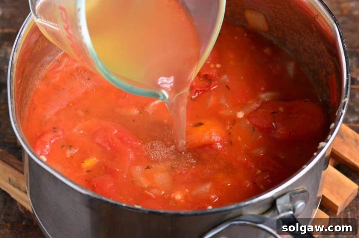 Pouring vegetable broth from a clear measuring cup into a simmering pot of vibrant red tomato basil soup.