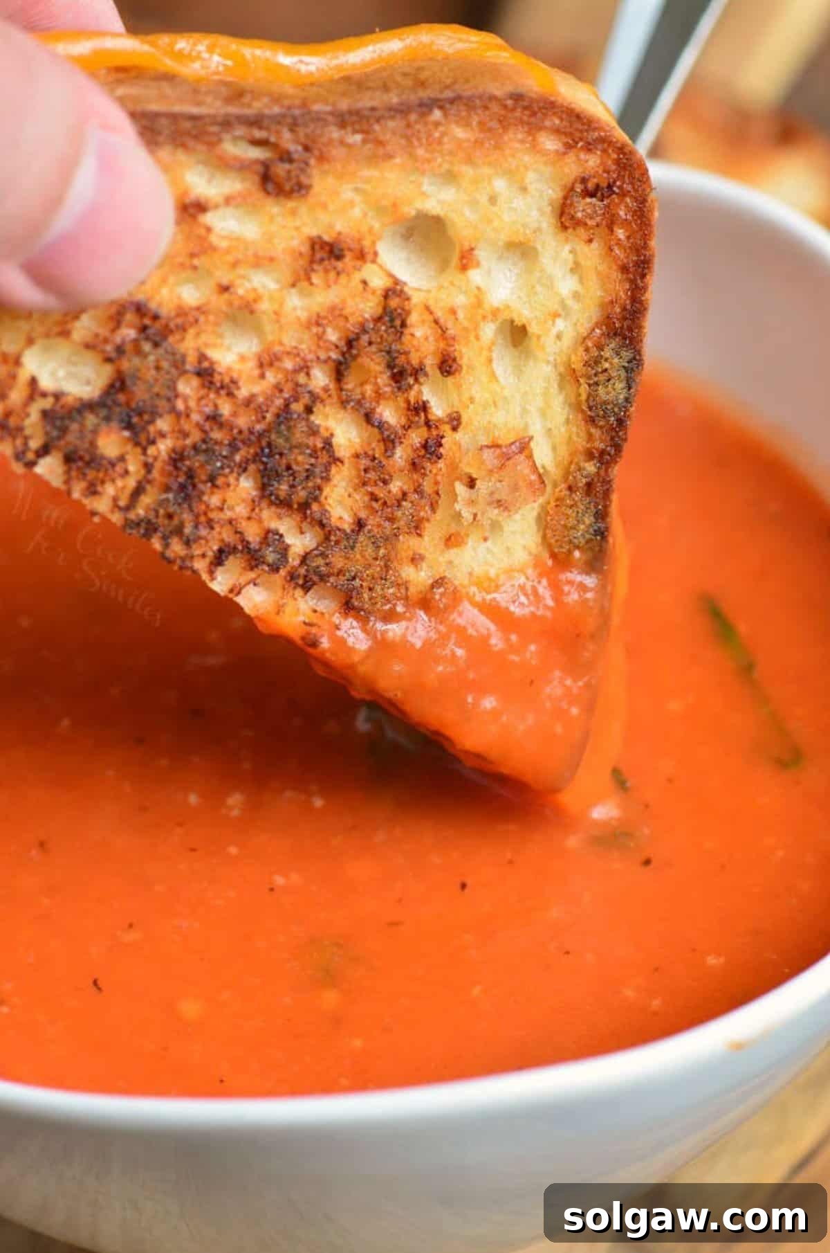 Close-up of tomato basil soup in a white bowl, with a corner of a grilled cheese sandwich being dipped into the rich, red soup.