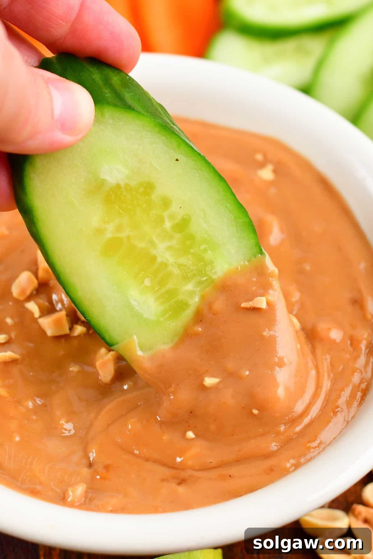 Closeup of dipping a fresh cucumber slice into creamy, homemade peanut sauce, showcasing its smooth texture and rich color.