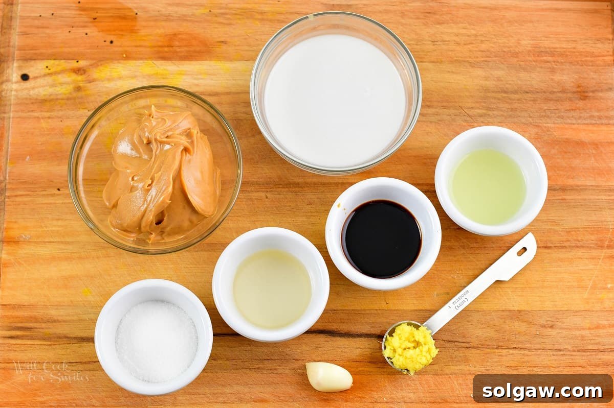 A selection of fresh ingredients for peanut sauce laid out on a wooden cutting board, including peanut butter, coconut milk, lime, garlic, and ginger.