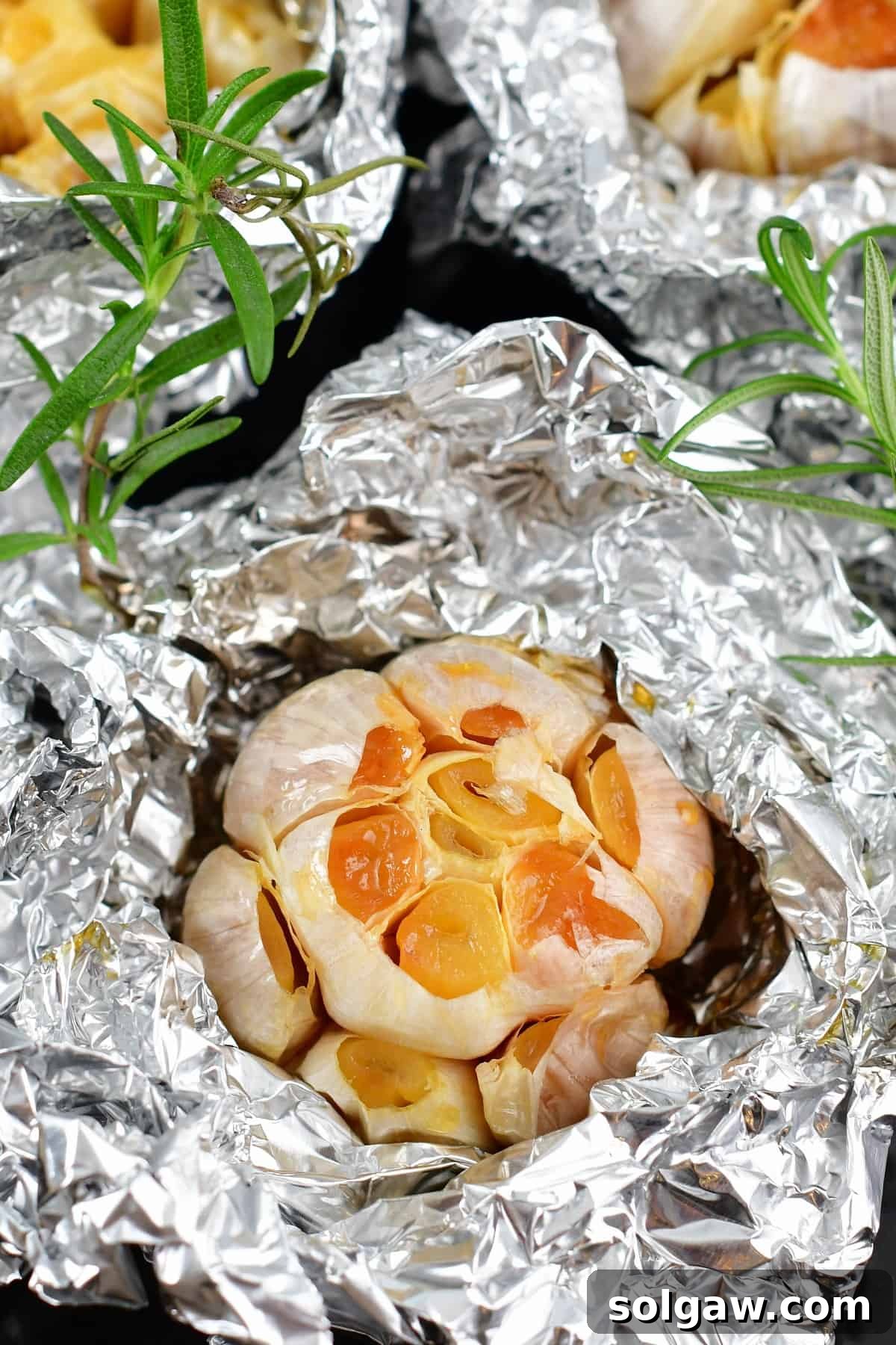 Sweet Mellow Roasted Garlic 2 Closeup of roasted garlic in foil with fresh rosemary sprigs, highlighting its golden-brown, tender texture.