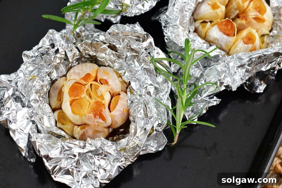 Sweet Mellow Roasted Garlic 5 Roasted garlic cloves still in foil on a baking sheet, accompanied by fresh rosemary sprigs, ready to be enjoyed.