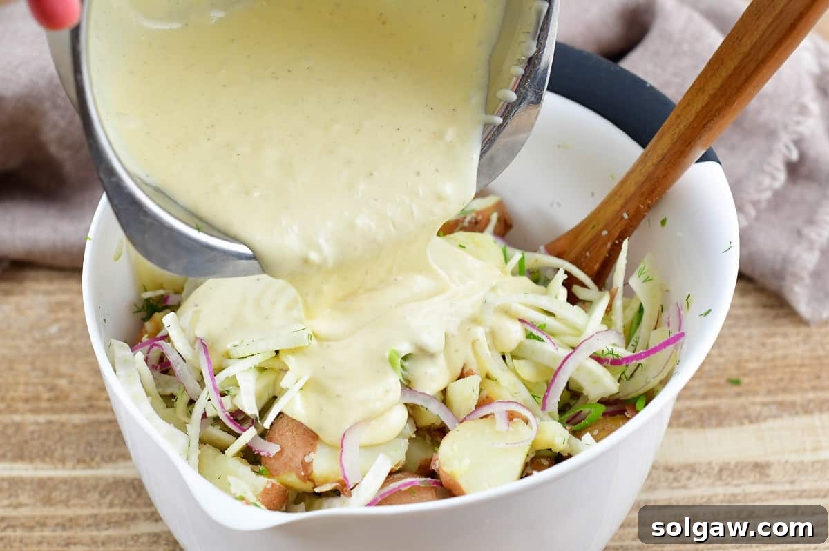 Adding the creamy potato salad dressing into a mixing bowl with cooked potatoes and other ingredients.