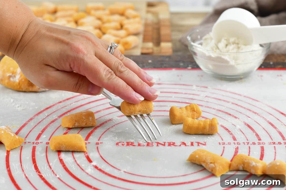A hand gently rolling a small piece of sweet potato gnocchi dough down the tines of a fork to create traditional ridges.