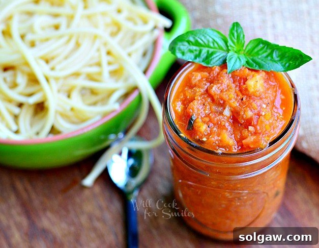 A bowl of freshly made Marinara sauce with a sprig of basil on the side, and a plate of pasta with sauce in the blurred background. The setup exudes a cozy, inviting meal.