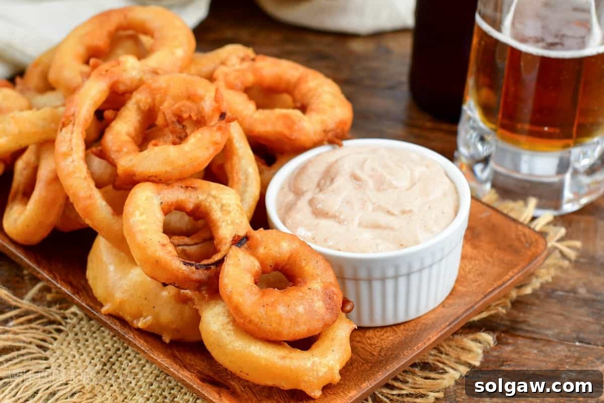 Beer Battered onion rings on a wood plate with a dipping sauce and a beer in the background.