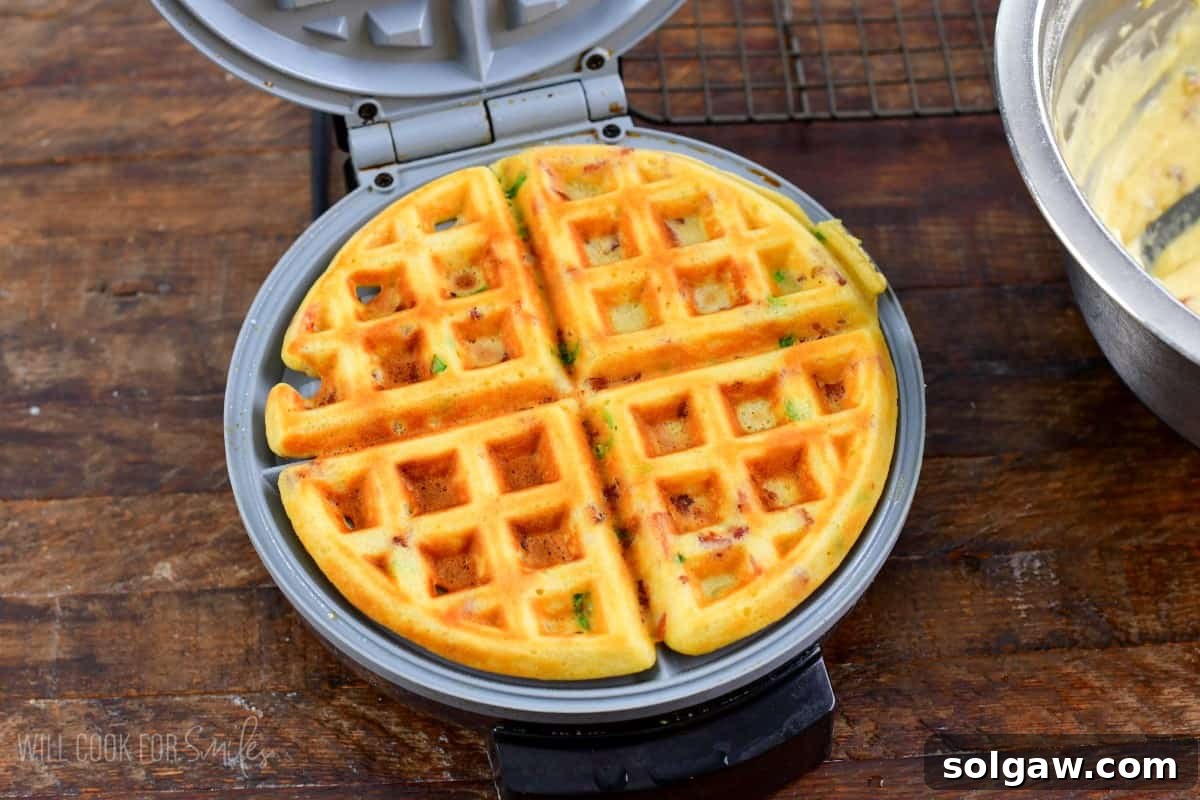 Cooked savory waffle in a waffle maker that is opened up and on a wood surface, steam gently rising.