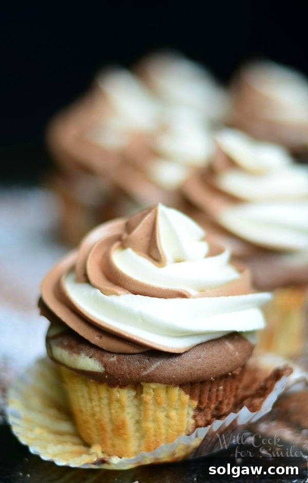Velvety Marble Cupcakes with Swirled Cream Cheese Frosting 8 An appetizing close-up of a Marble Cupcake with its liner peeled away, resting on a black countertop sprinkled with powdered sugar, inviting a bite.